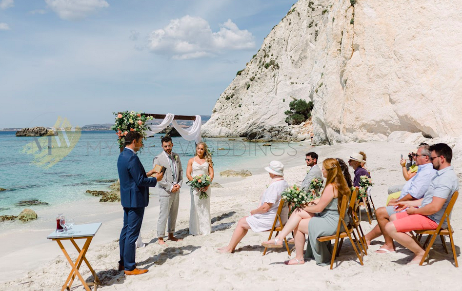 Ein Hochzeitspaar am Strand steht unter einem Blumenbogen und hört einem Redner zu. Davor sitzen Menschen auf Stühlen.