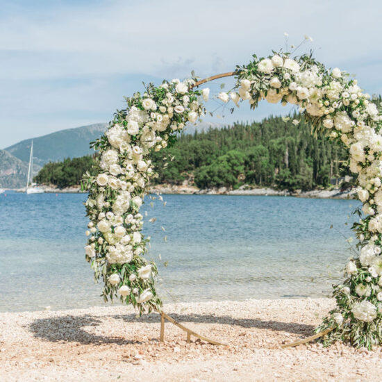 Runde Hochzeitsbogen aus Metall mit grün-weißen Blumen am Strand in Griechenland mit Segelbooten im Hintergrund