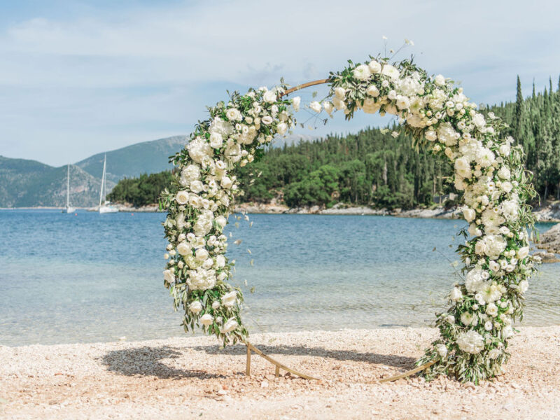Runde Hochzeitsbogen aus Metall mit grün-weißen Blumen am Strand in Griechenland mit Segelbooten im Hintergrund