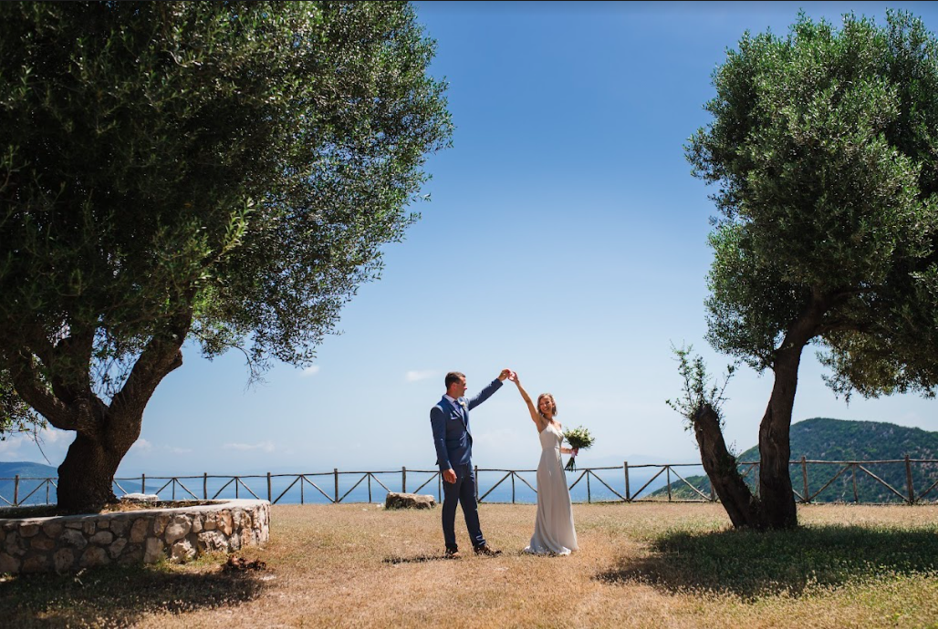 Braut und Bräutigam stehen auf einem Staubplatz mit Meerblick im Hintergrund und heben die Hände