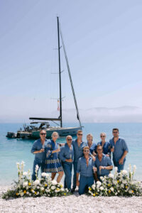 Personen am Strand mit Blumenschmuck, Segelboot im Hintergrund